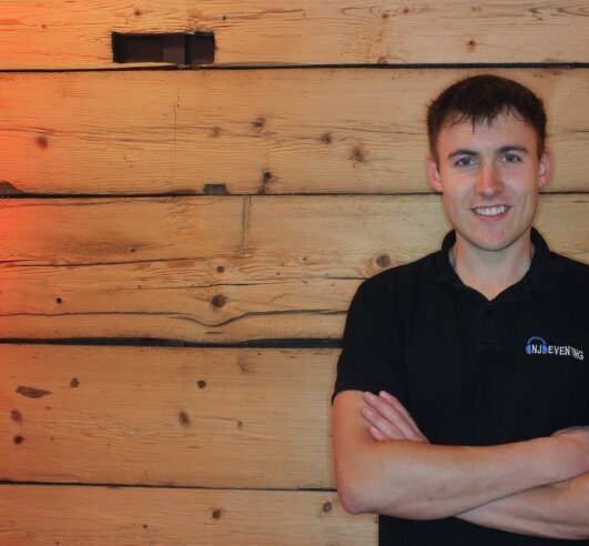 nico_japes_hochzeits_dj_nj_eventing_kirchhundem_sauerland Nico Japes, professioneller Hochzeits-DJ und Inhaber von NJ-Eventing aus Kirchhundem. Er lächelt im Marken-Shirt vor einer Holzwand. Ihr Experte für Eventtechnik im Kreis Olpe und Sauerland.